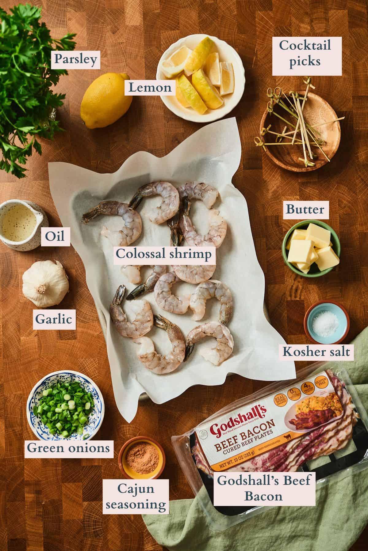 Labeled ingredients for Cajun beef bacon wrapped shrimp including colossal shrimp, Godshall's beef bacon, Cajun seasoning, butter, garlic, lemon, parsley, green onions, kosher salt, oil, and bamboo cocktail picks on a wooden board.