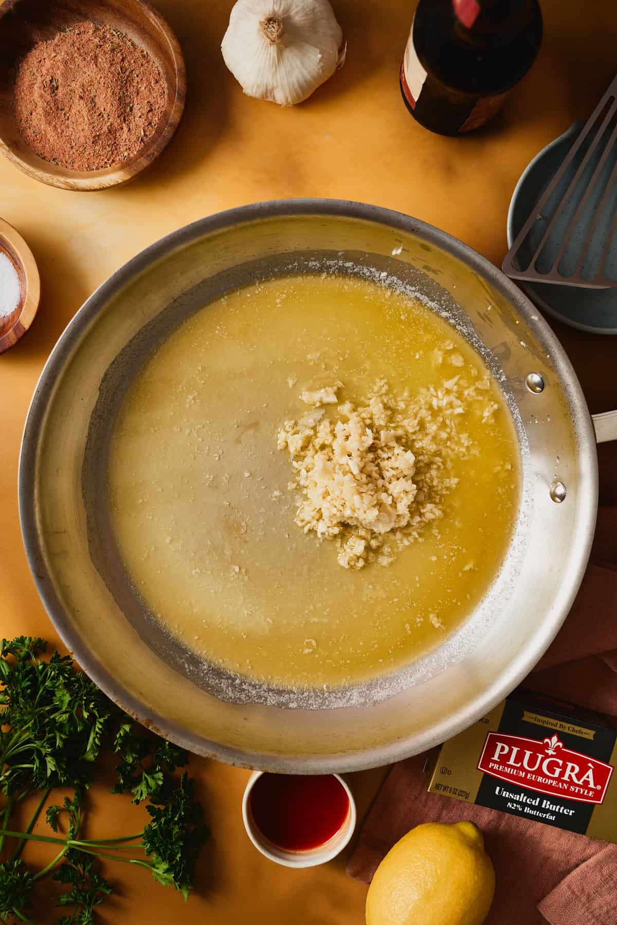 Garlic softening in melted Plugrà butter in a skillet, pale gold and fragrant, a fish spatula stirring it through.