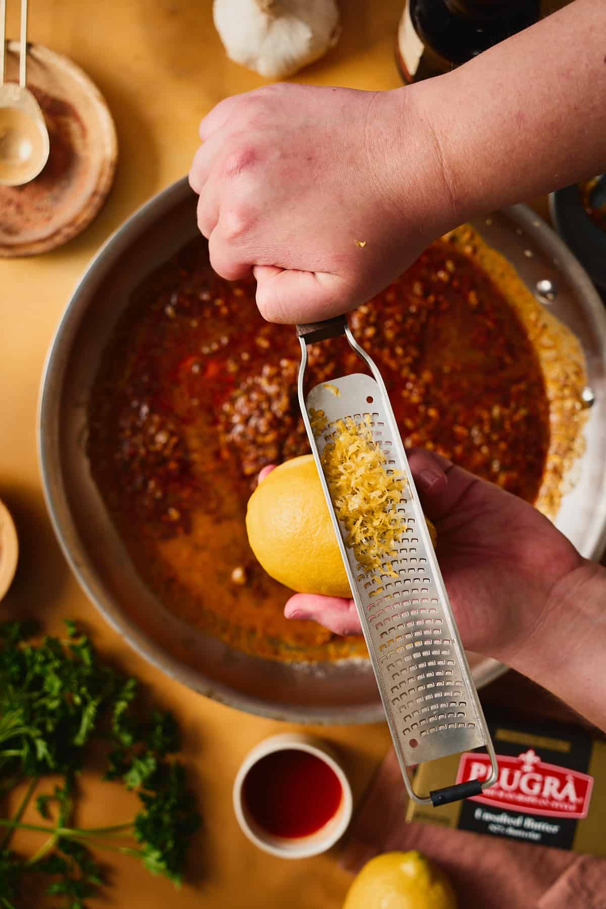 A hand grating fresh lemon zest from a microplane into a skillet of garlic butter sauce.