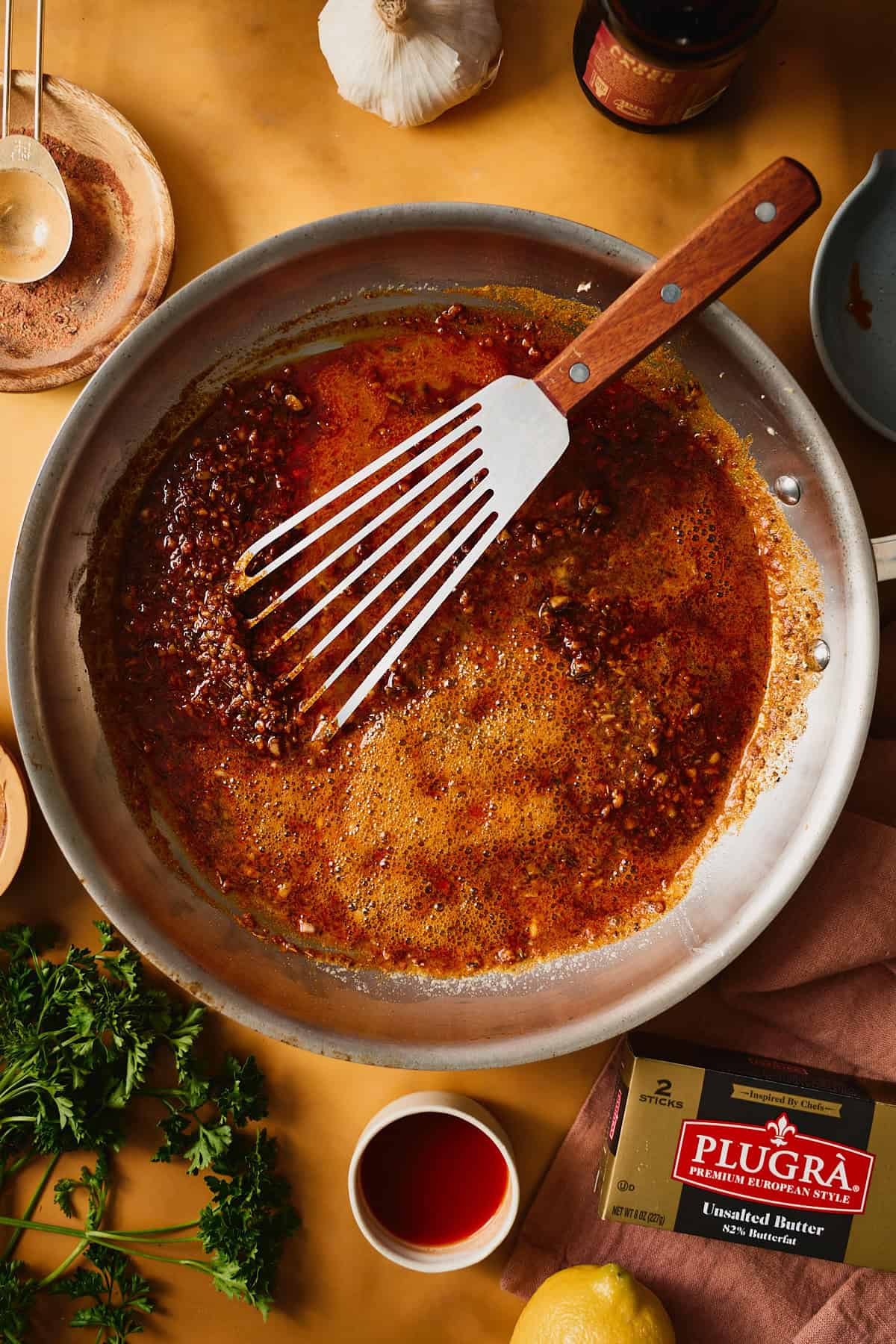 Garlic butter sauce in a skillet after the beer goes in, deep red with minced garlic visible, a fish spatula resting in the pan.
