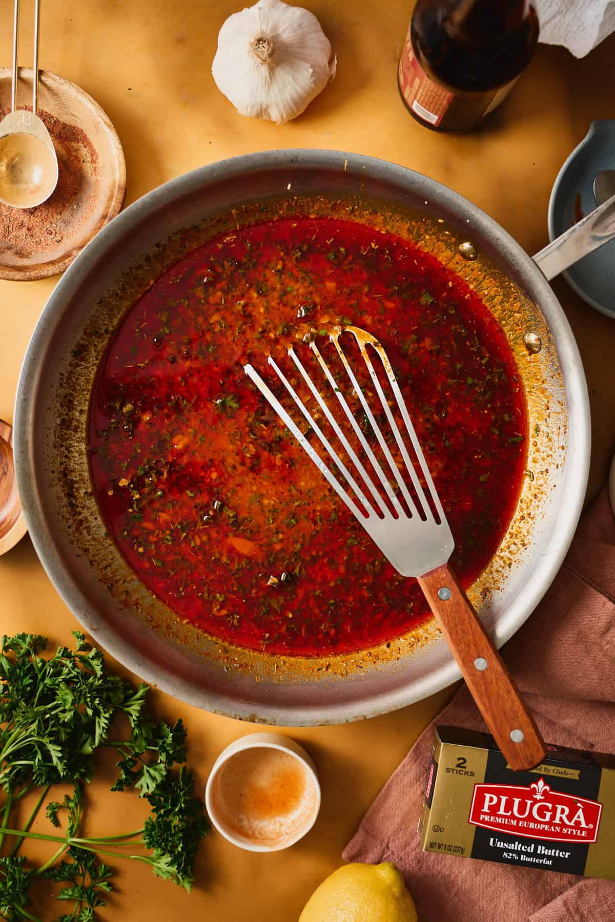 Finished Cajun garlic butter seafood boil sauce in a stainless skillet, glossy red-orange and flecked with parsley, a fish spatula resting in the pan.