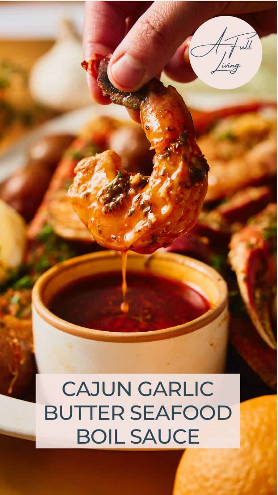 A hand dipping a saucy shrimp into a bowl of deep red Cajun garlic butter seafood boil sauce, with crab legs and potatoes on the platter behind it and a text overlay reading Cajun Garlic Butter Seafood Boil Sauce.