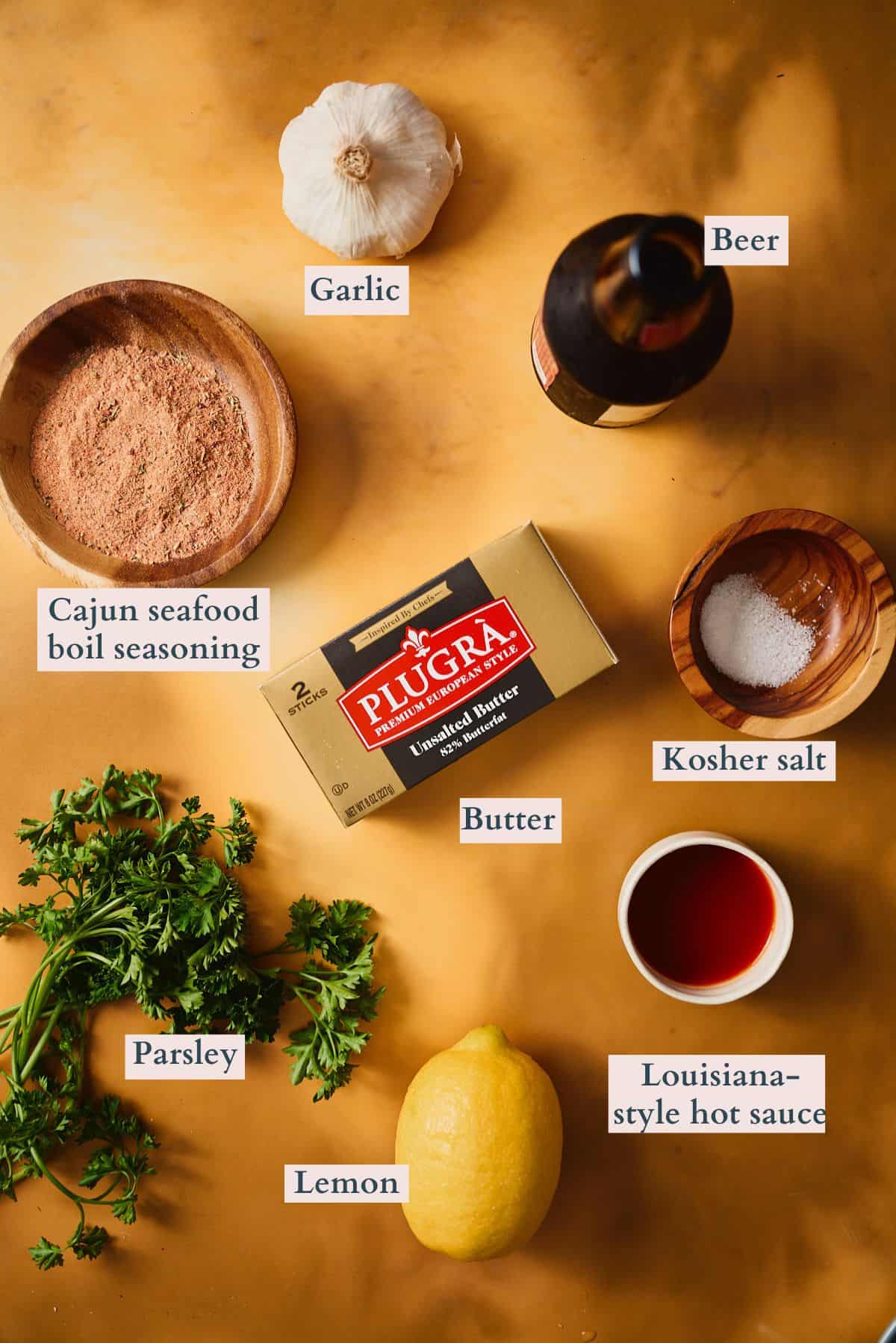 Labeled ingredients for garlic butter seafood boil sauce on a warm terracotta background, including Plugrà European-style butter, garlic, Cajun seafood boil seasoning, kosher salt, lemon, Louisiana-style hot sauce, parsley, and beer.