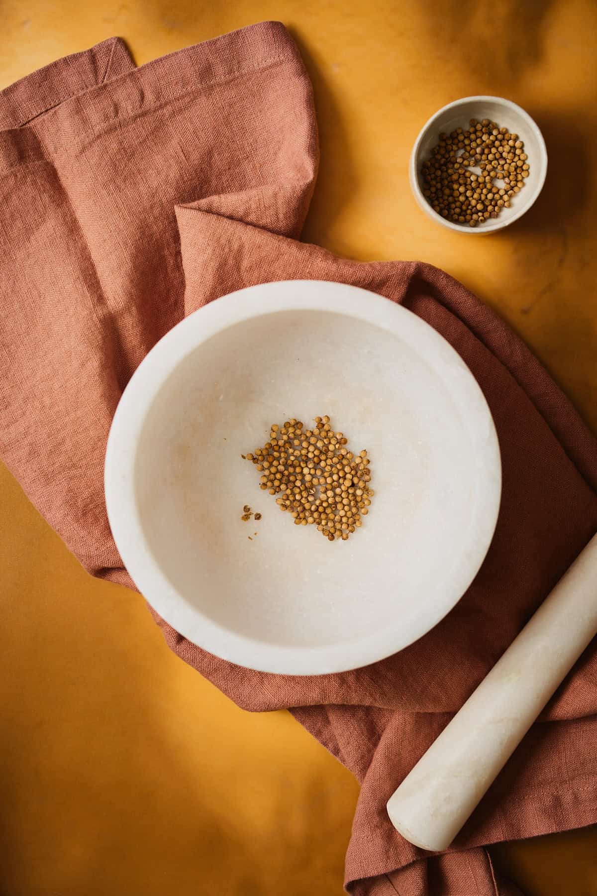 Whole coriander seeds resting in a white marble mortar with the pestle alongside.