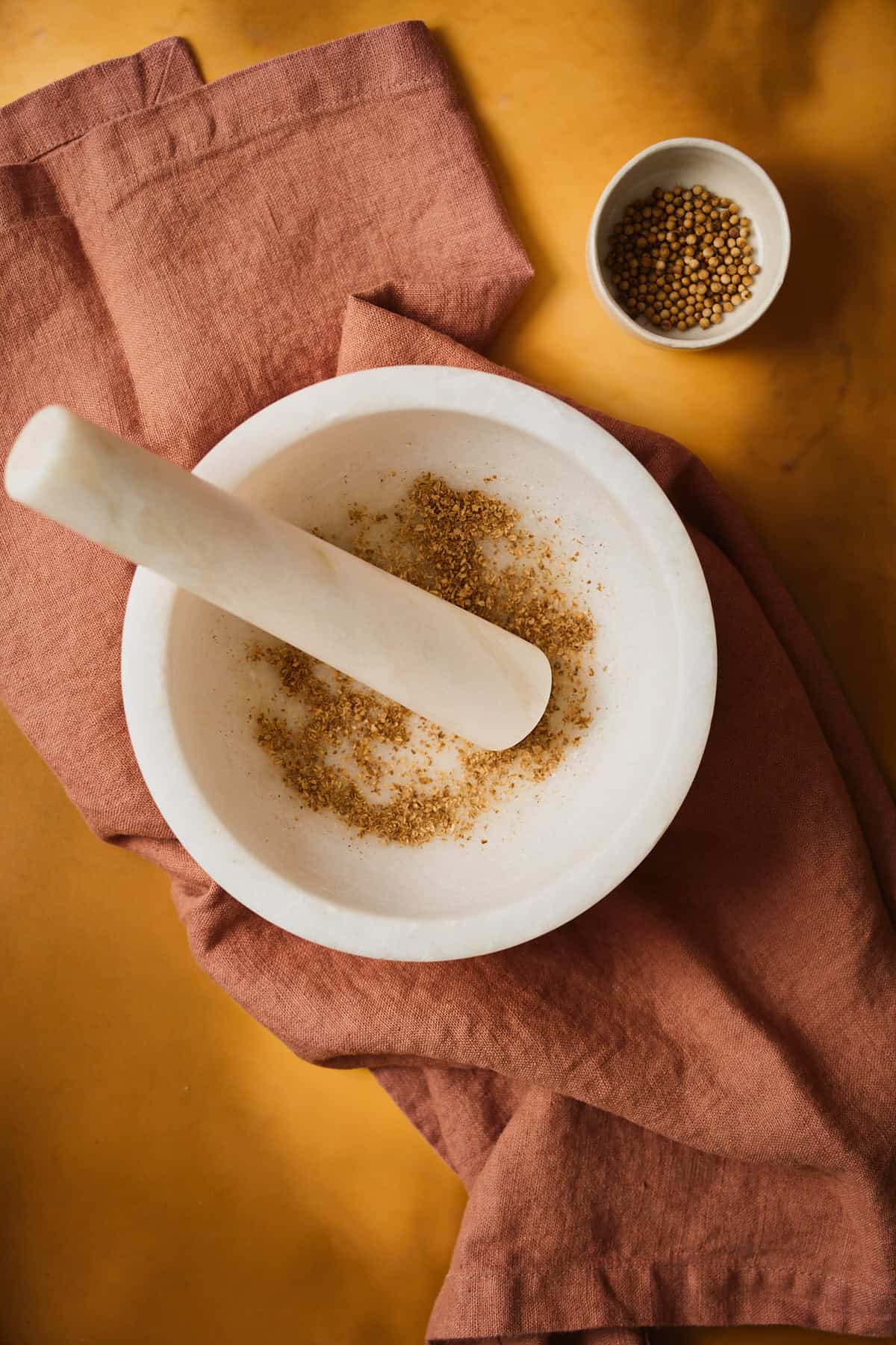 Coriander seeds partway ground to a coarse powder in a marble mortar, pestle resting inside.