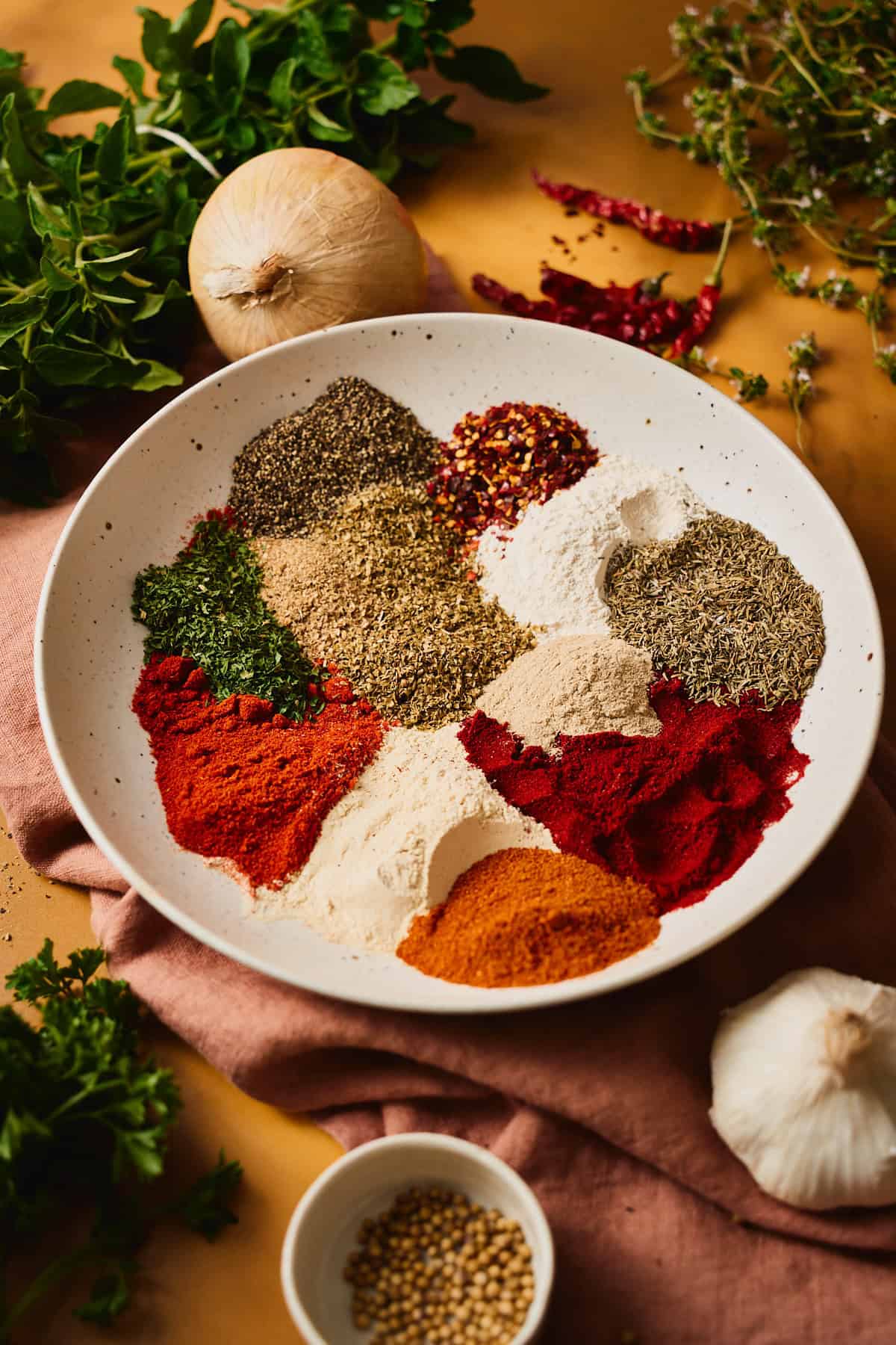Individual mounds of paprika, cayenne, dried herbs, garlic and onion powder, and peppercorns laid out in a speckled bowl before mixing at a 45-degree angle surrounded by aromatics.