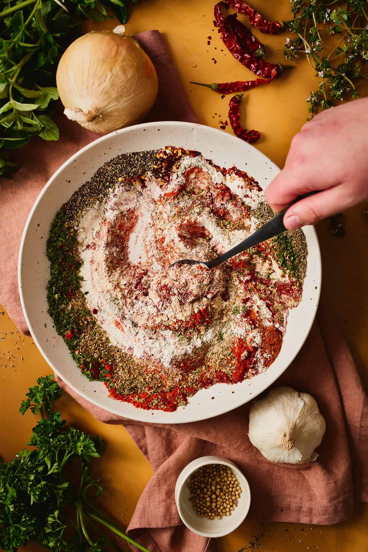 A hand stirring the half-blended Cajun seasoning, paprika and herbs streaking together in a speckled bowl.