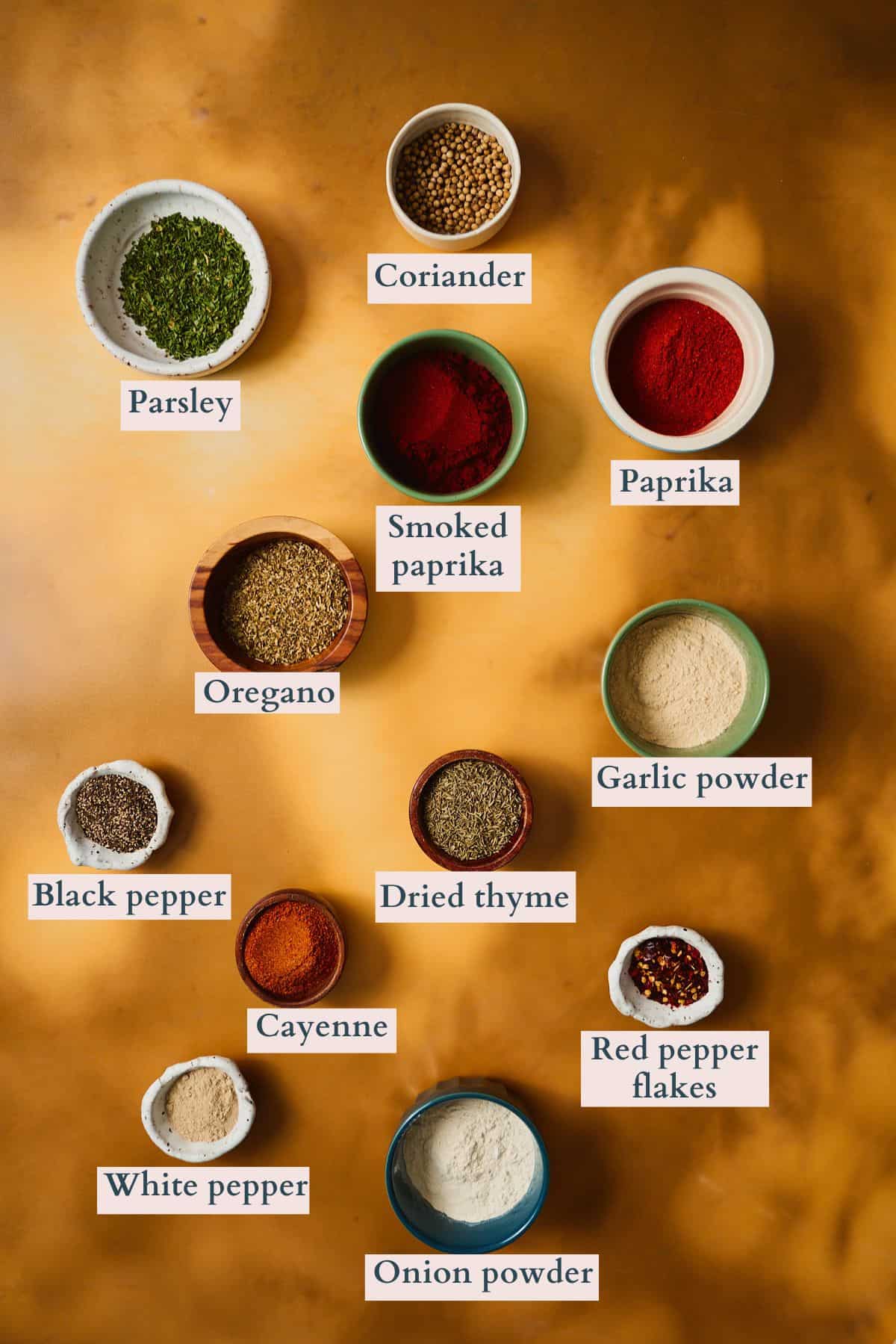 Labeled overhead flat lay of Cajun seafood boil seasoning ingredients in small bowls, including parsley, coriander, smoked paprika, paprika, oregano, garlic powder, black pepper, dried thyme, cayenne, red pepper flakes, white pepper, and onion powder.
