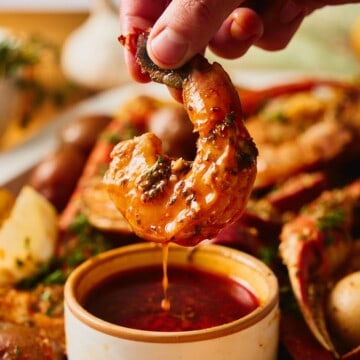 A hand lifting a glossy, sauce-coated shrimp out of a small bowl of Cajun garlic butter seafood boil sauce, a drip falling back into the bowl, with snow crab legs, red potatoes, and sausage on the loaded platter behind it.