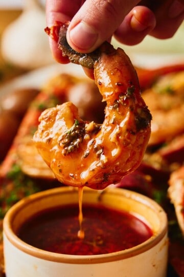 A hand lifting a glossy, sauce-coated shrimp out of a small bowl of Cajun garlic butter seafood boil sauce, a drip falling back into the bowl, with snow crab legs, red potatoes, and sausage on the loaded platter behind it.