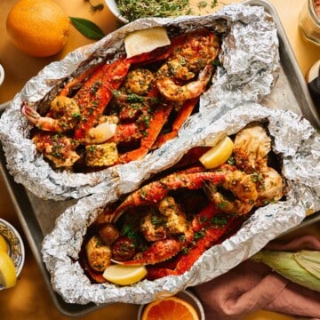 Two open foil packets of Cajun garlic butter seafood boil with snow crab legs, jumbo shrimp, sliced sausage, corn, and baby potatoes on a metal sheet pan, garnished with parsley and fresh lemon wedges.