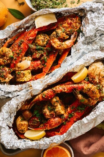 Two open foil packets of Cajun garlic butter seafood boil with snow crab legs, jumbo shrimp, sliced sausage, corn, and baby potatoes on a metal sheet pan, garnished with parsley and fresh lemon wedges.