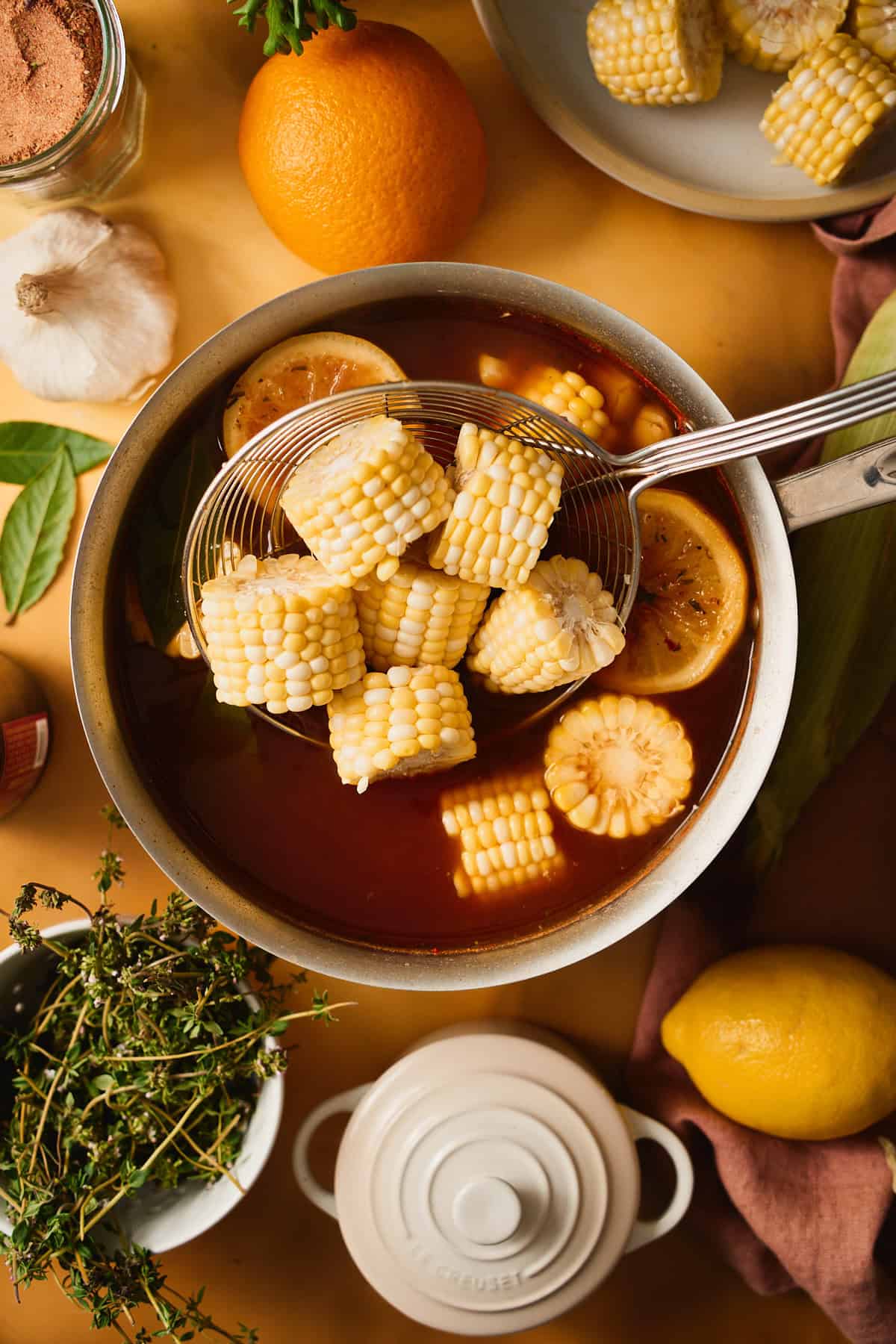 Sweet corn cobbettes in a spider strainer being lifted into seasoned par-boil water with a charred lemon half floating alongside.