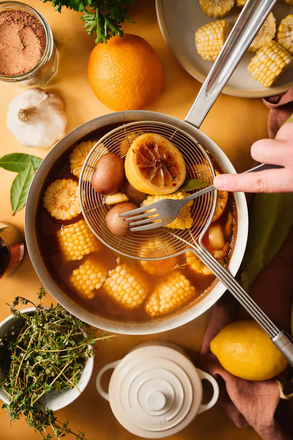 Hand pulling out corn and potatoes from seasoned boiling water and piercing a potato with a fork.