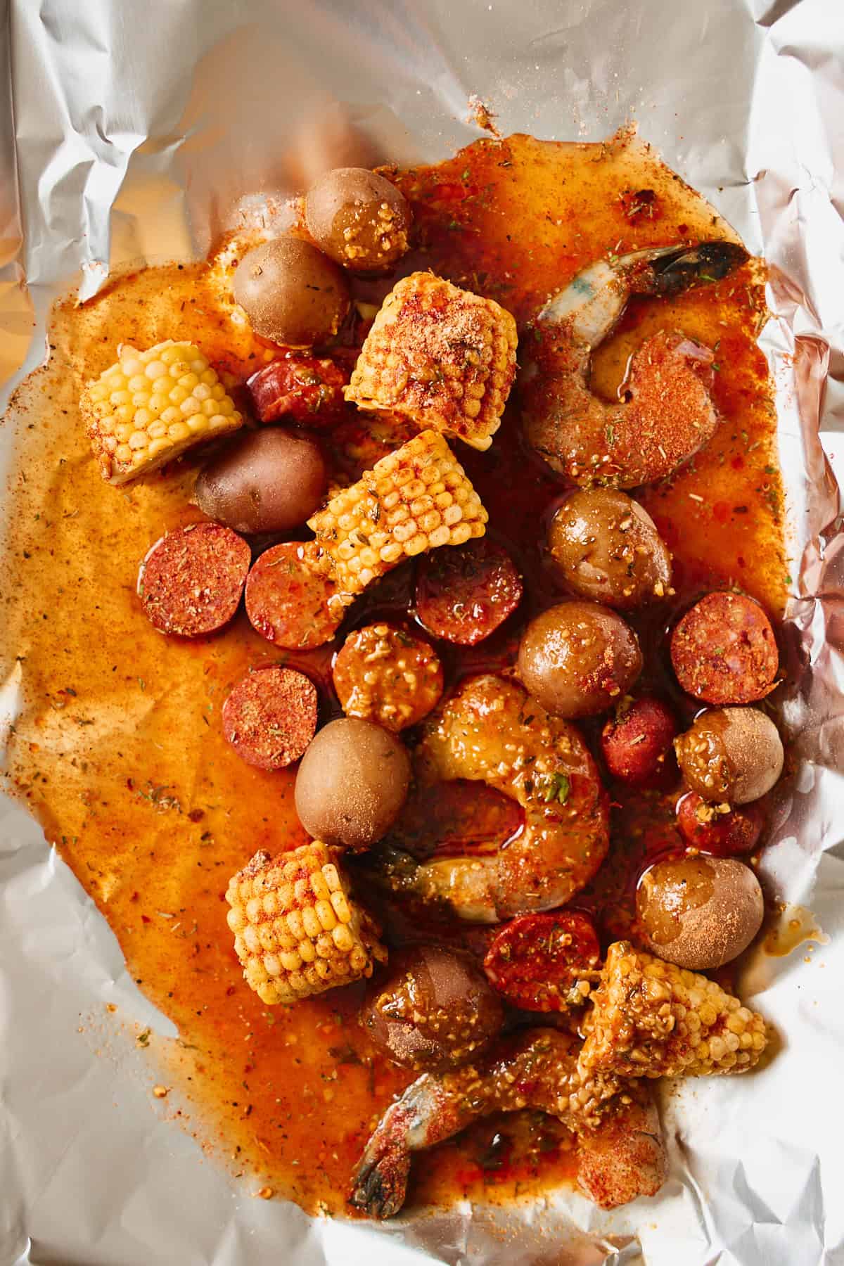 Raw seasoned shrimp pressed down into the buttery sauce pool at the bottom of a foil packet with potatoes, sausage, and corn.
