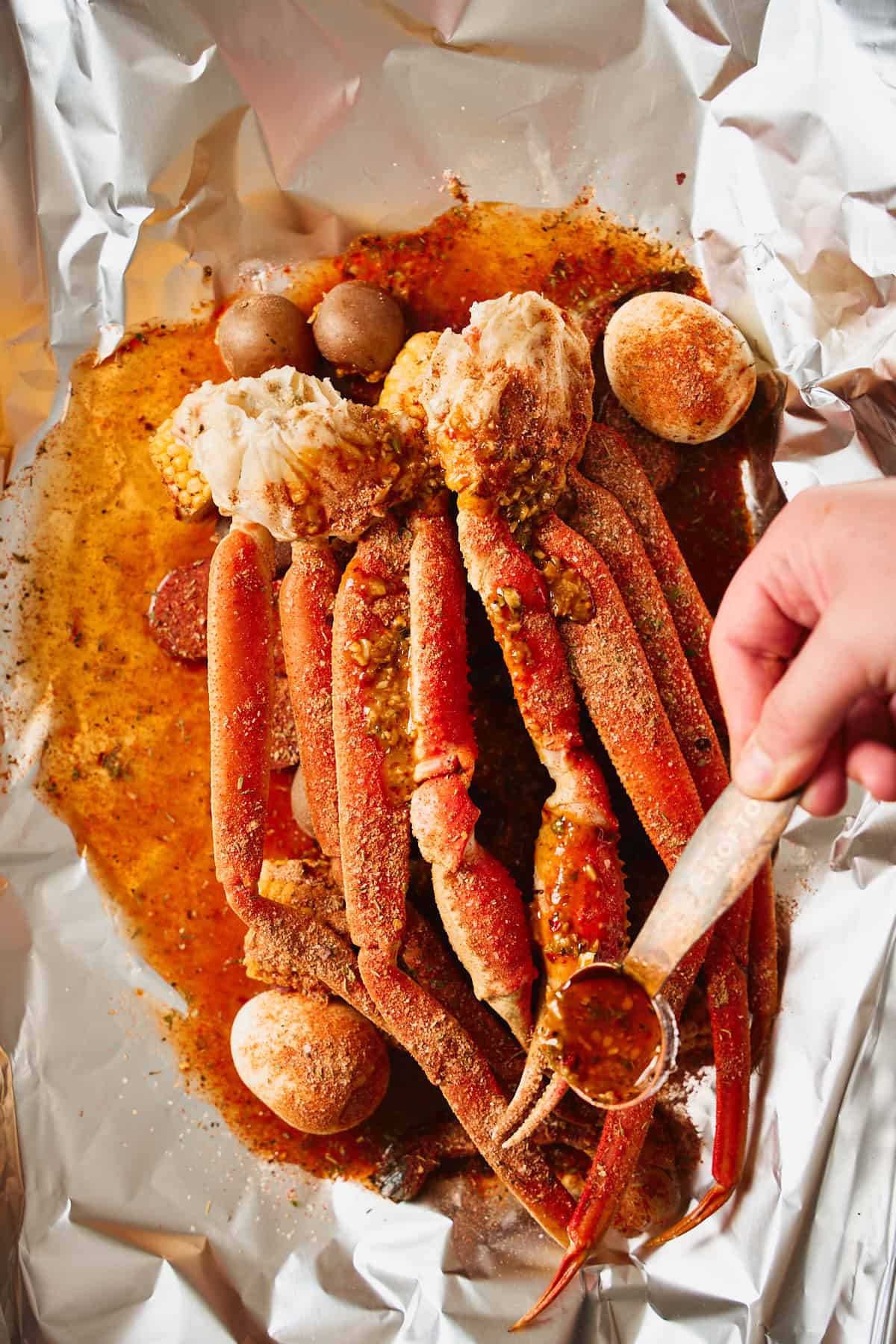 A ladle drizzling garlic butter sauce over an assembled foil packet of snow crab, corn, potatoes, and seasoning before sealing.
