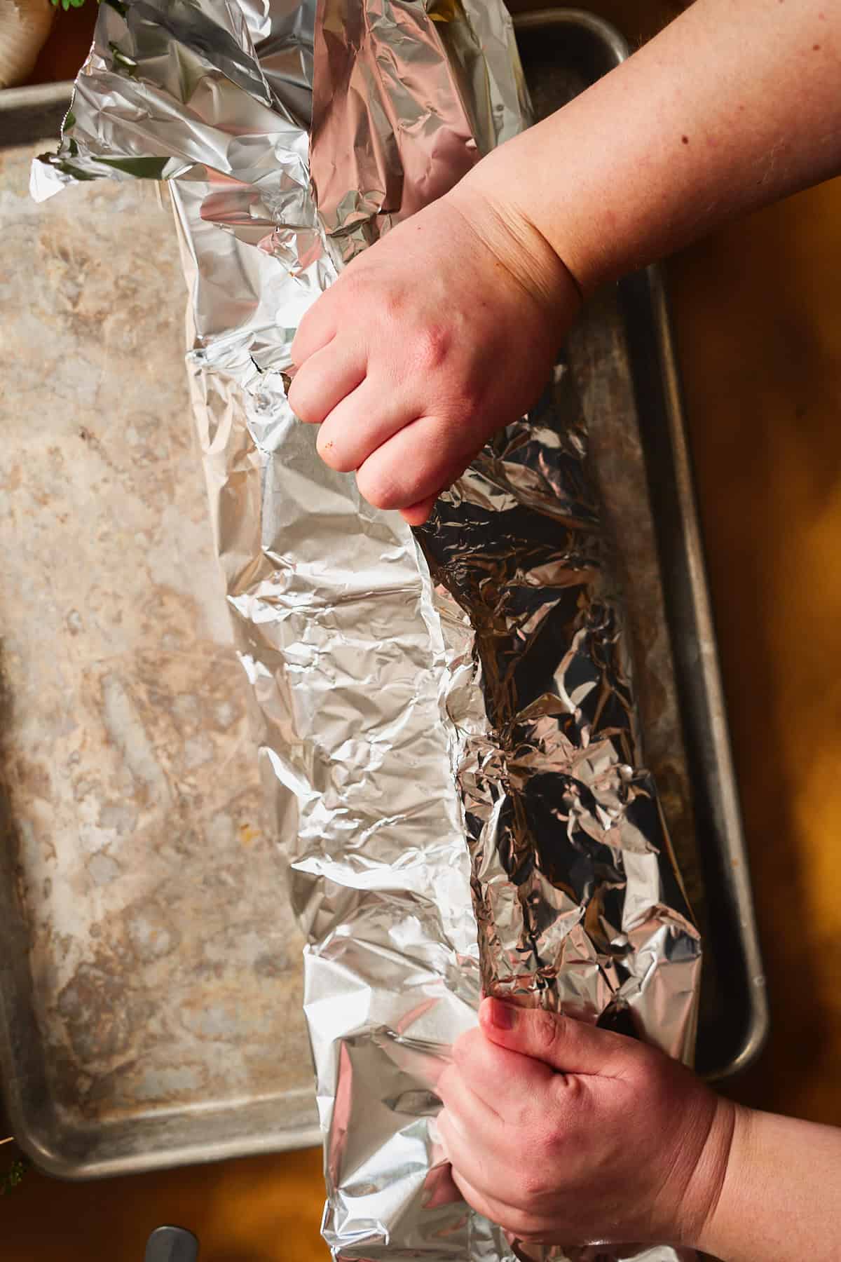 Hands crimping the long sides of a foil packet together over a sheet pan to seal in the seafood boil.