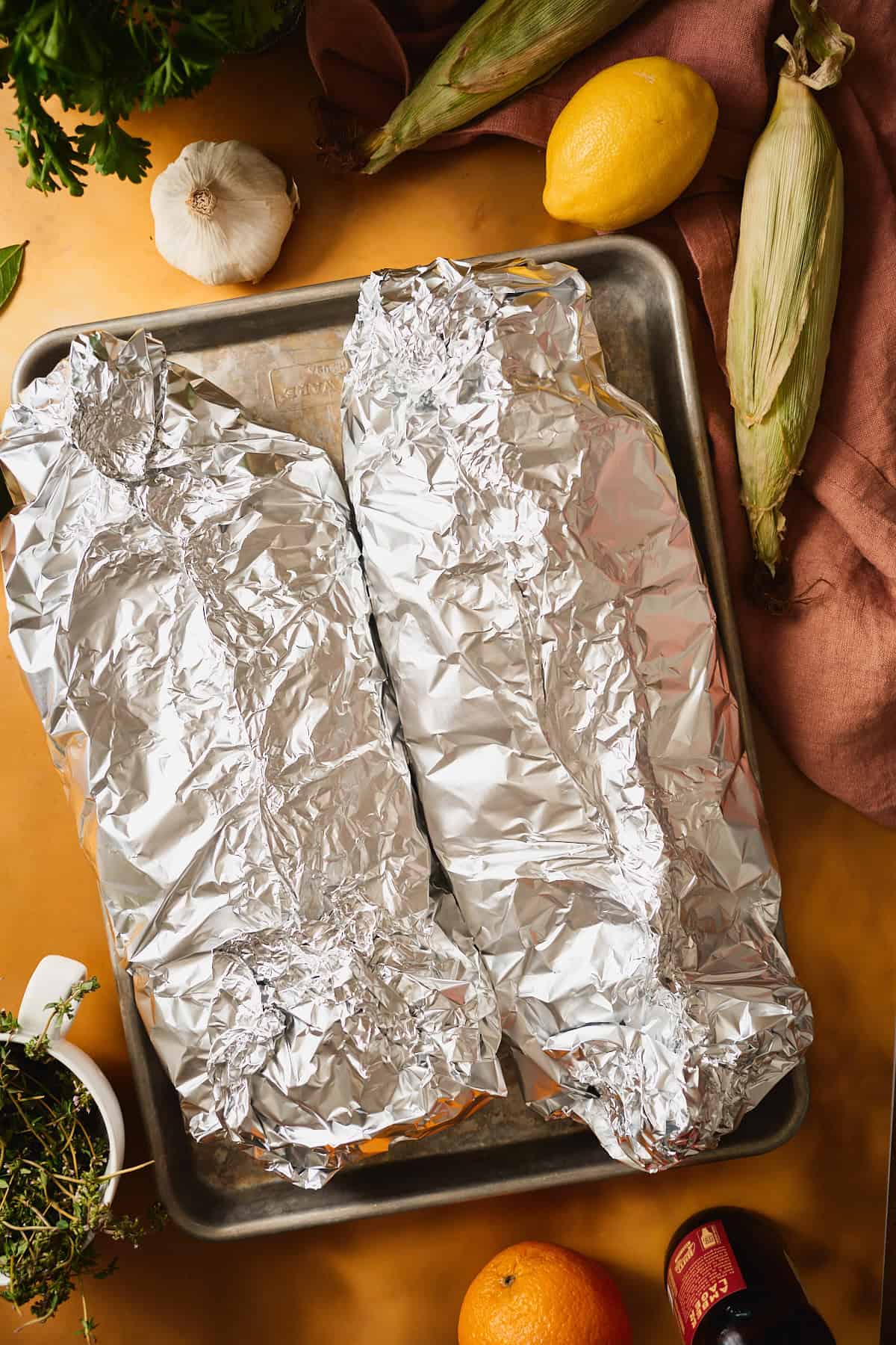 Two sealed foil packets on a metal sheet pan ready for the oven, with corn, lemon, and garlic styled alongside.