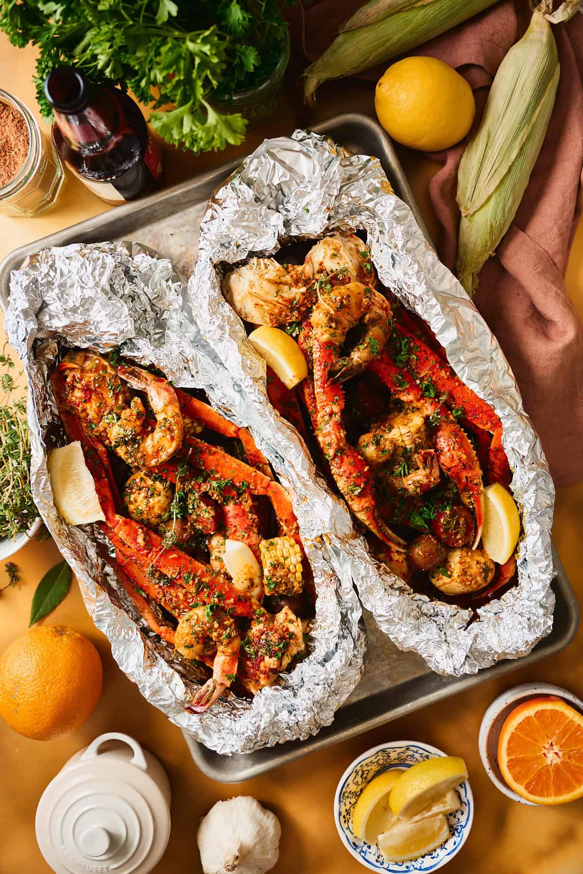 Two open foil packets of Cajun garlic butter seafood boil with snow crab, shrimp, sausage, corn, and potatoes on a sheet pan, garnished with parsley and lemon wedges.