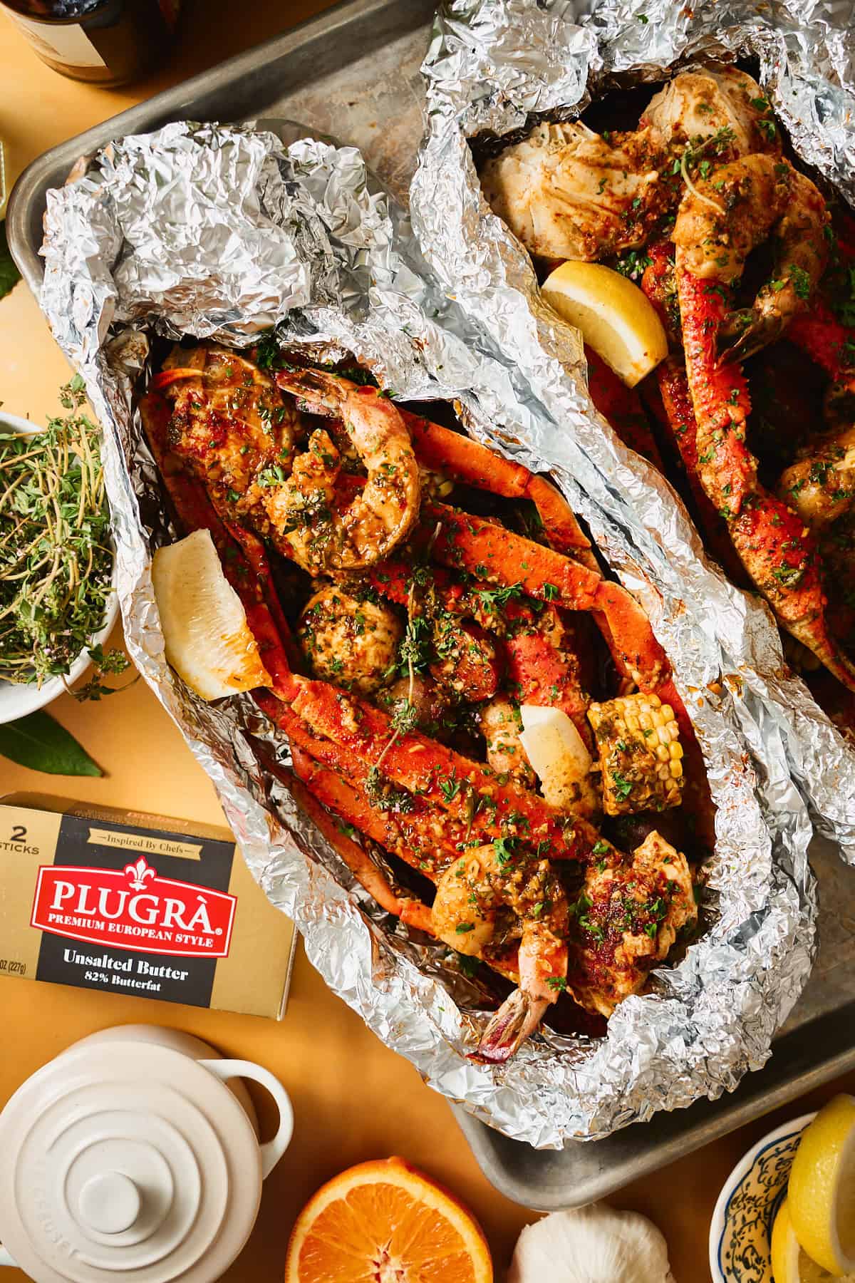 Two open foil packets of Cajun garlic butter seafood boil with snow crab, shrimp, corn, and potatoes on a sheet pan, a box of Plugrà butter and fresh herbs in the frame.
