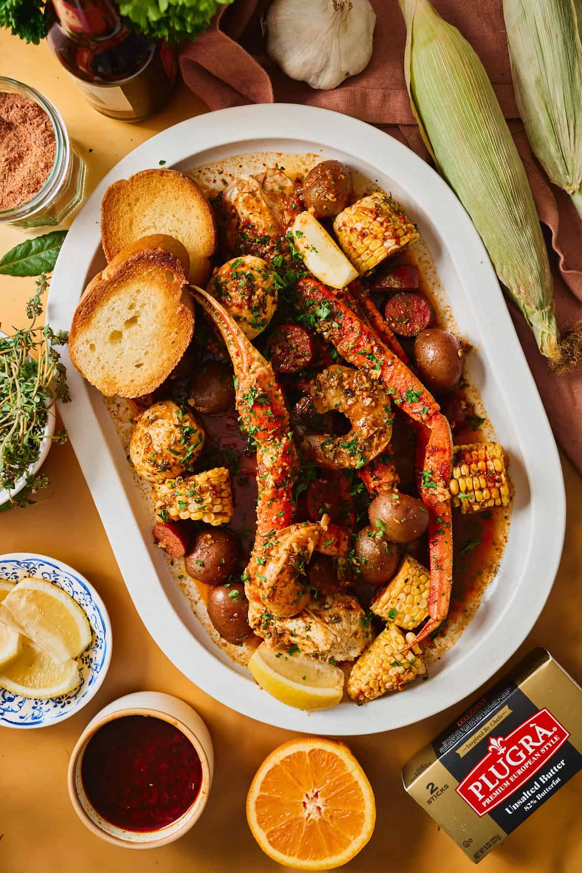 Cajun garlic butter seafood boil with snow crab, shrimp, andouille, corn, and potatoes plated on a white oval platter with toasted bread, a ramekin of dipping sauce, and a box of Plugrà butter alongside.