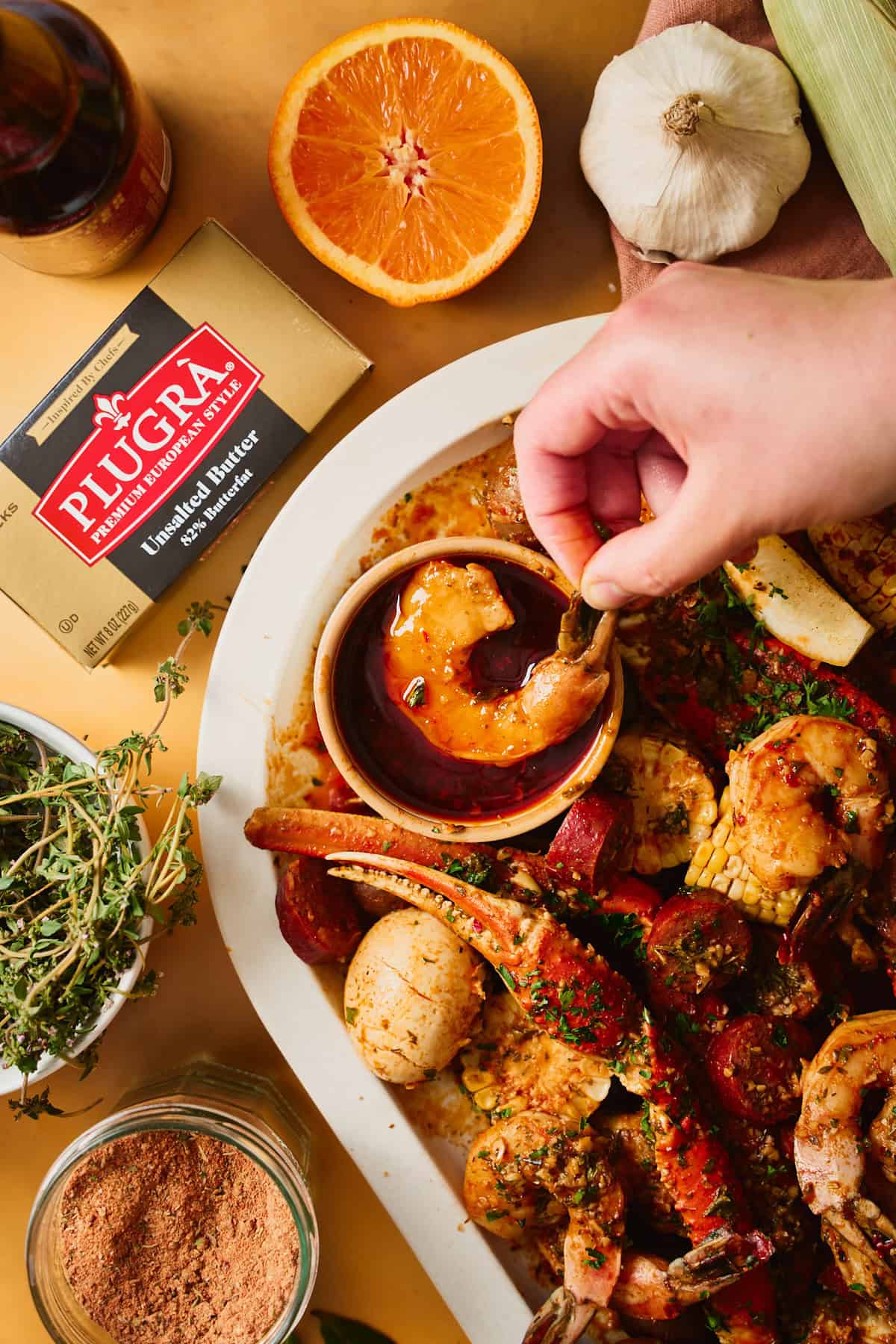 A hand dipping a saucy shrimp into a bowl of garlic butter seafood boil sauce, with a Plugrà European-style butter box and a jar of seasoning in frame beside a loaded seafood platter.