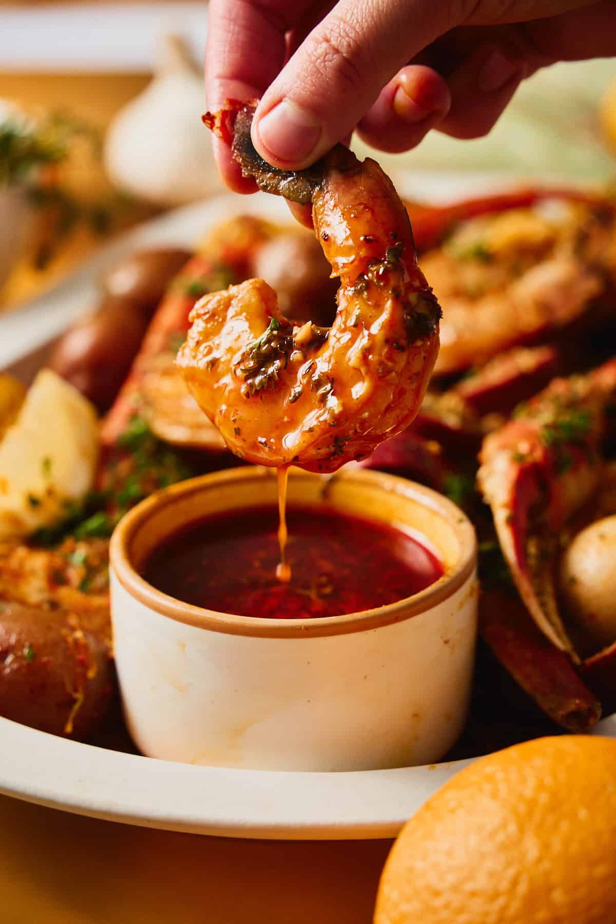 Hand dipping a butter-glazed jumbo shrimp into a ramekin of Cajun garlic butter sauce, the sauce dripping back into the dish, with crab, potatoes, and corn behind.