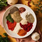 Individual mounds of smoked and sweet paprika, cayenne, dried herbs, garlic and onion powder, peppercorns, and red pepper flakes arranged in a speckled bowl, ready to mix into Cajun seafood boil seasoning.