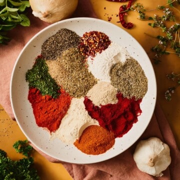 Individual mounds of smoked and sweet paprika, cayenne, dried herbs, garlic and onion powder, peppercorns, and red pepper flakes arranged in a speckled bowl, ready to mix into Cajun seafood boil seasoning.