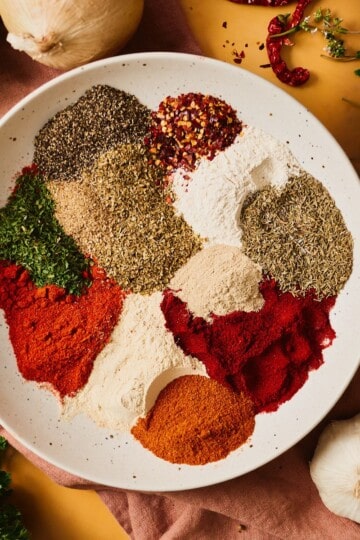 Individual mounds of smoked and sweet paprika, cayenne, dried herbs, garlic and onion powder, peppercorns, and red pepper flakes arranged in a speckled bowl, ready to mix into Cajun seafood boil seasoning.