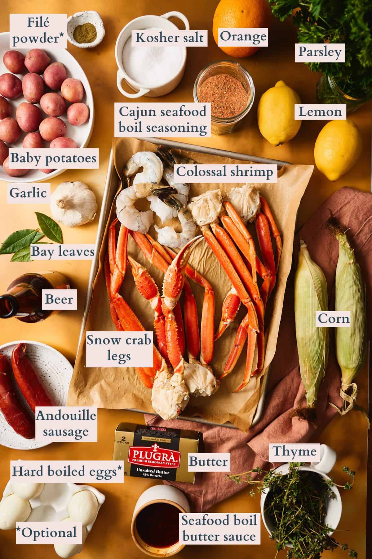 Labeled ingredients for Cajun garlic butter shrimp and crab foil packets, including snow crab legs, colossal shrimp, andouille sausage, baby potatoes, corn, garlic, lemon, orange, hard boiled eggs, Cajun seafood boil seasoning, filé powder, beer, and Plugrà butter.