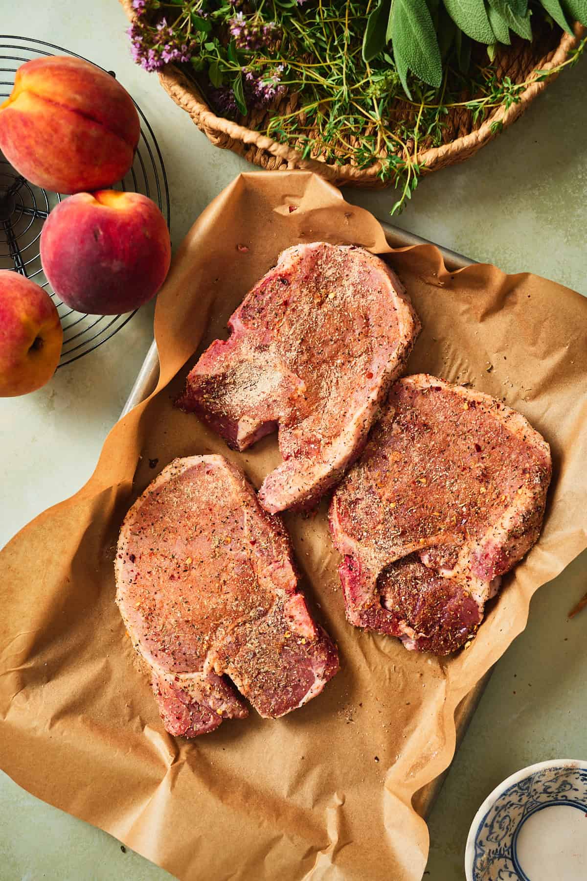 Three seasoned bone-in pork chops on parchment with fresh peaches and garden herbs.