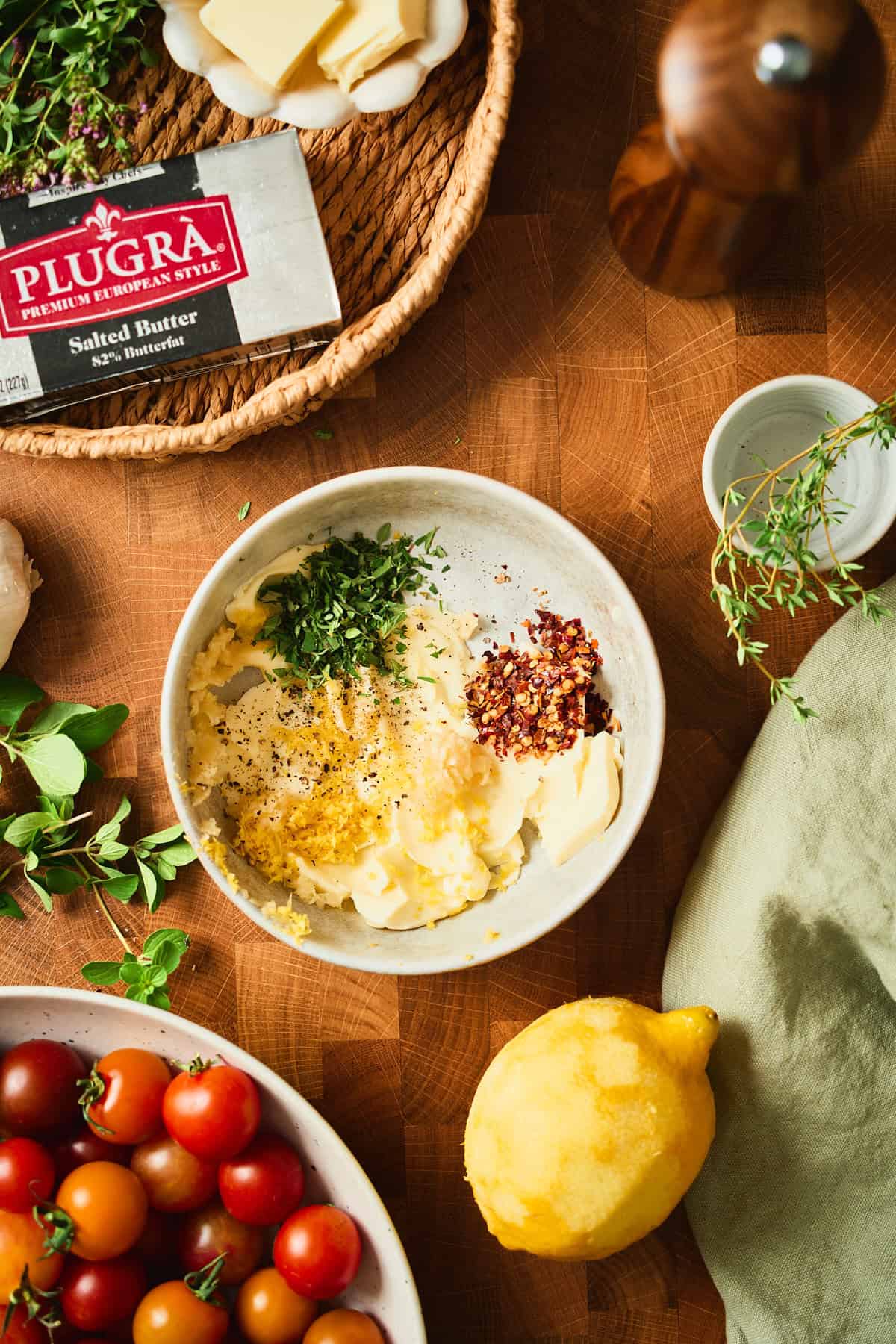 Softened Plugrà butter in a bowl with grated garlic, lemon zest, fresh thyme, oregano, red pepper flakes, and black pepper for a tomato galette.