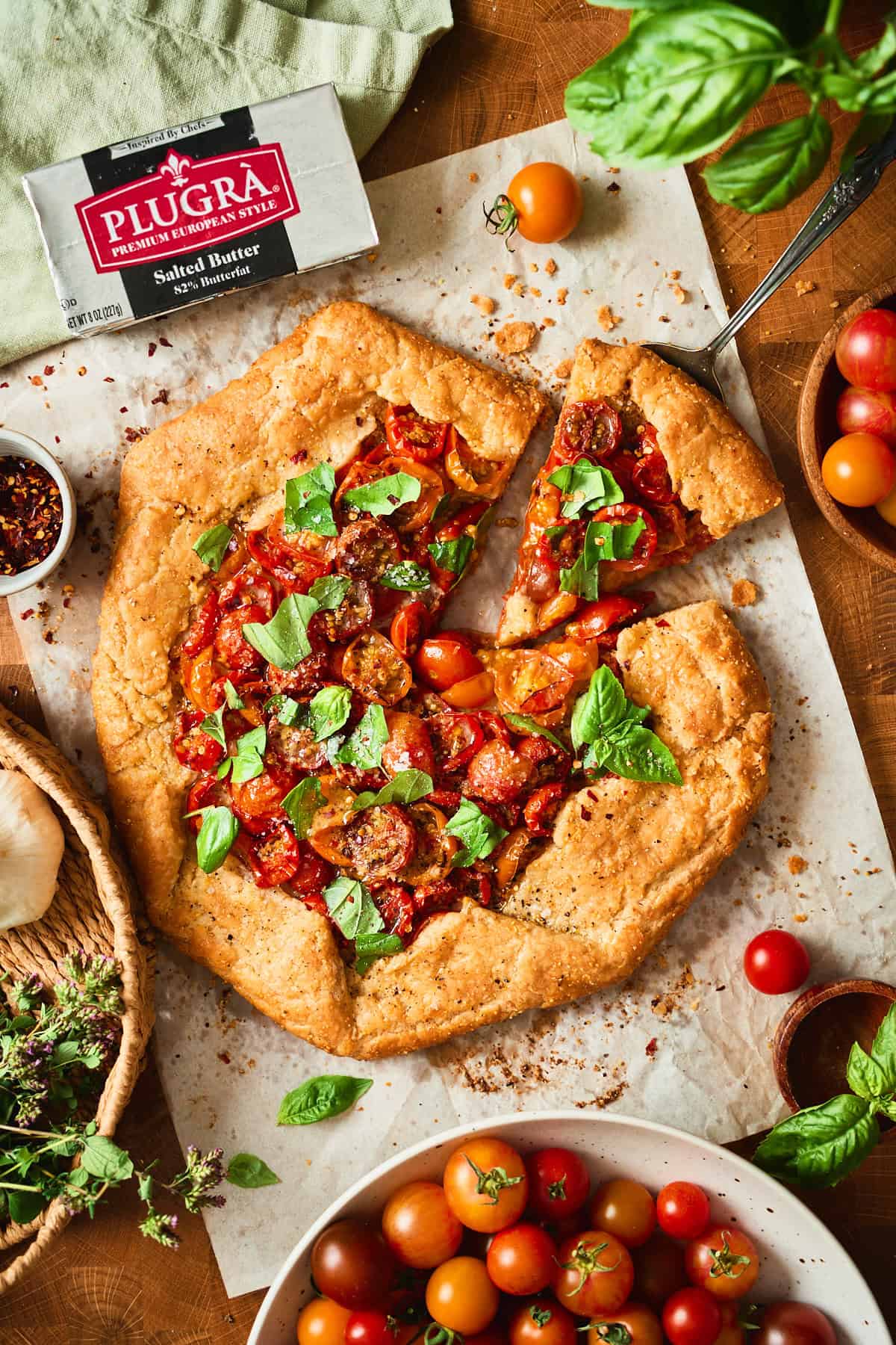 Sliced cherry tomato galette on parchment next to a package of Plugrà salted European-style butter, with fresh basil and cherry tomatoes.