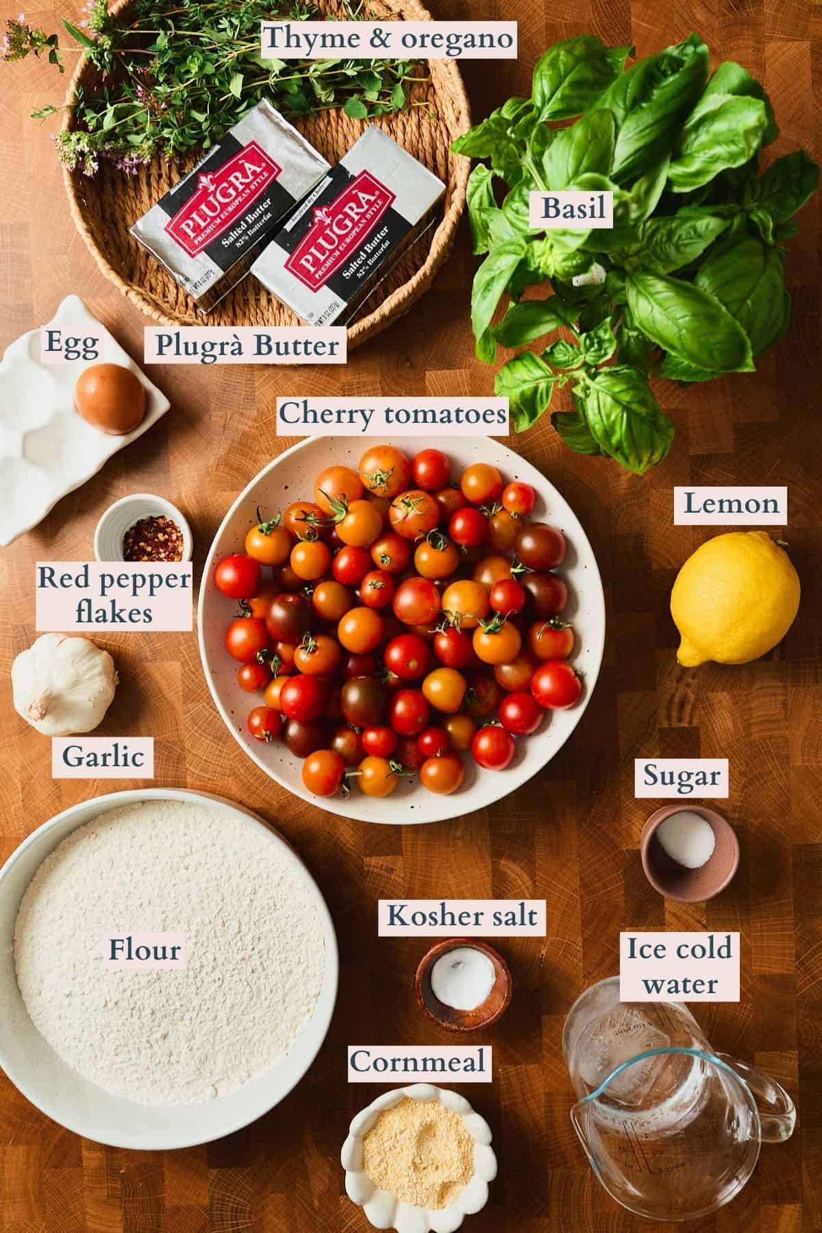 Labeled ingredients for a tomato galette recipe including cherry tomatoes, Plugrà salted butter, flour, cornmeal, basil, thyme, oregano, garlic, lemon, egg, red pepper flakes, sugar, kosher salt, and ice water.