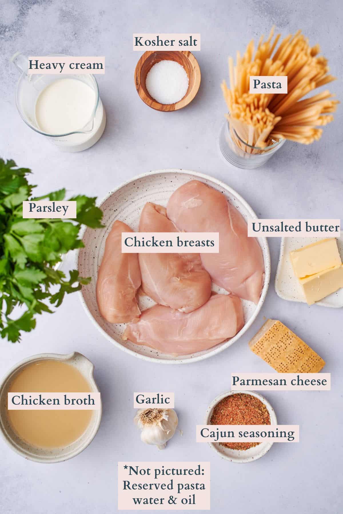 Ingredients to make cajun chicken alfredo laid out on a table including chicken breasts, Parmesan, parsley, garlic, heavy cream, chicken broth, cajun seasoning, salt,  butter, and pasta.