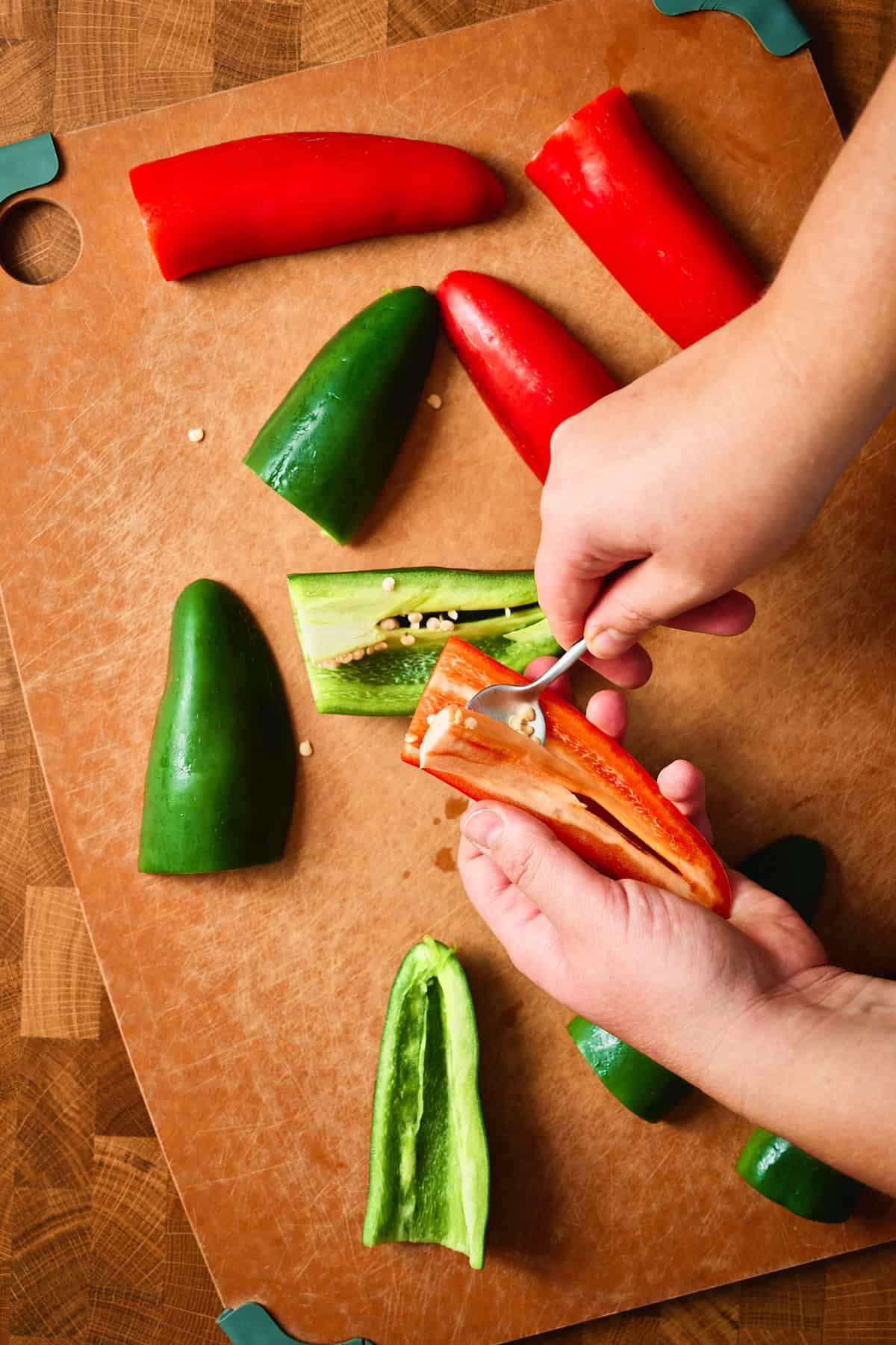 Hands scooping seeds from halved red and green jalapeños with a small spoon on a cutting board.