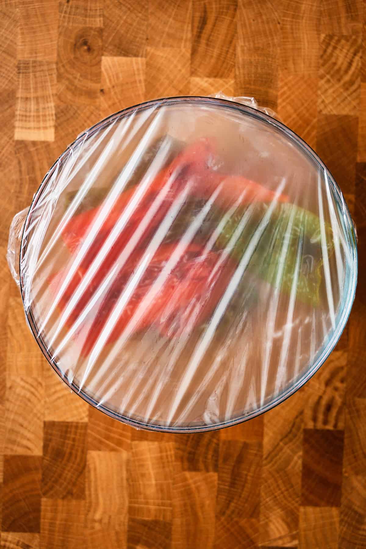 A glass bowl of broiled jalapeños covered tightly with plastic wrap to steam.