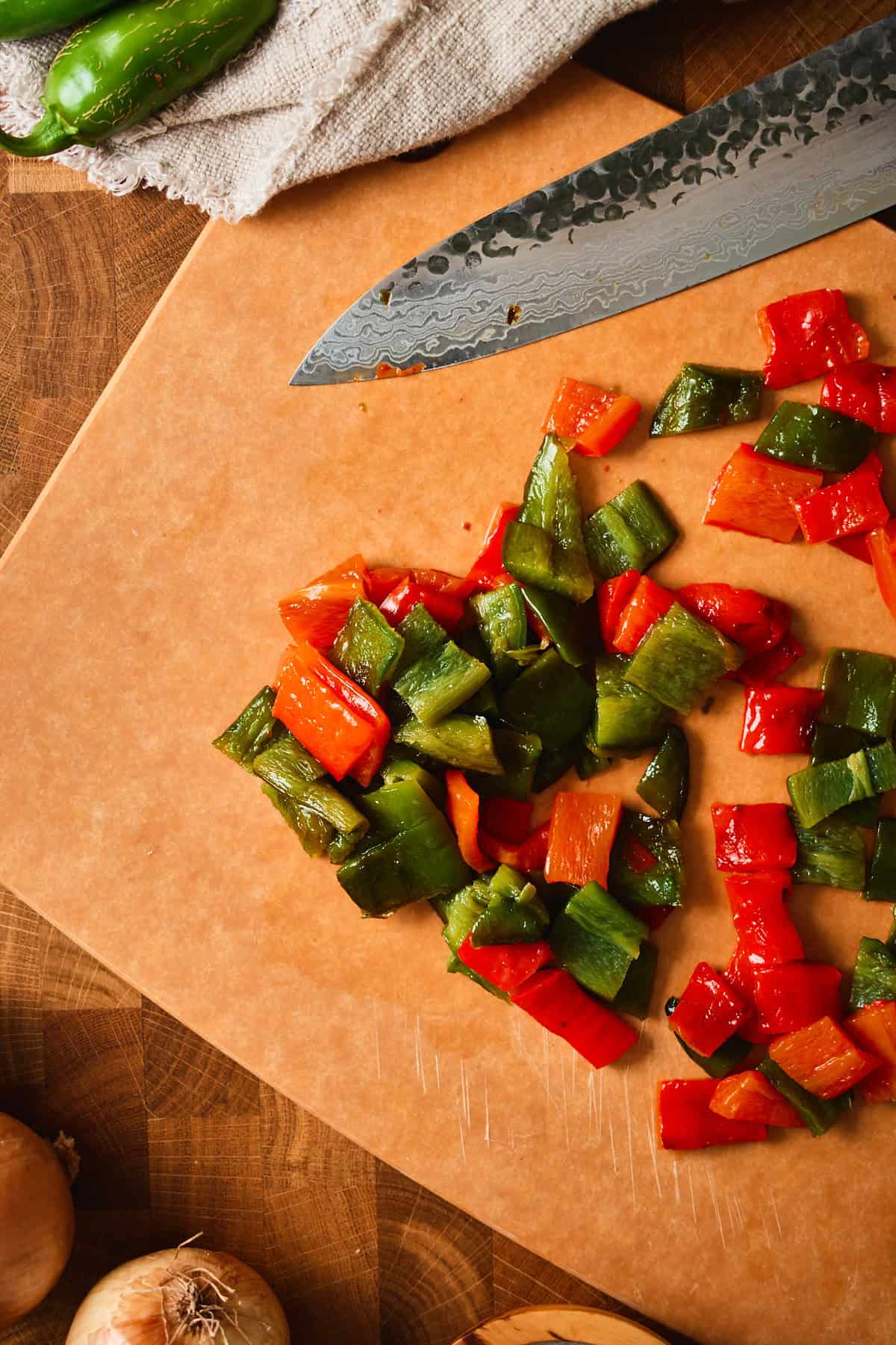 Peeled roasted red and green jalapeños chopped on a cutting board next to a chef's knife.
