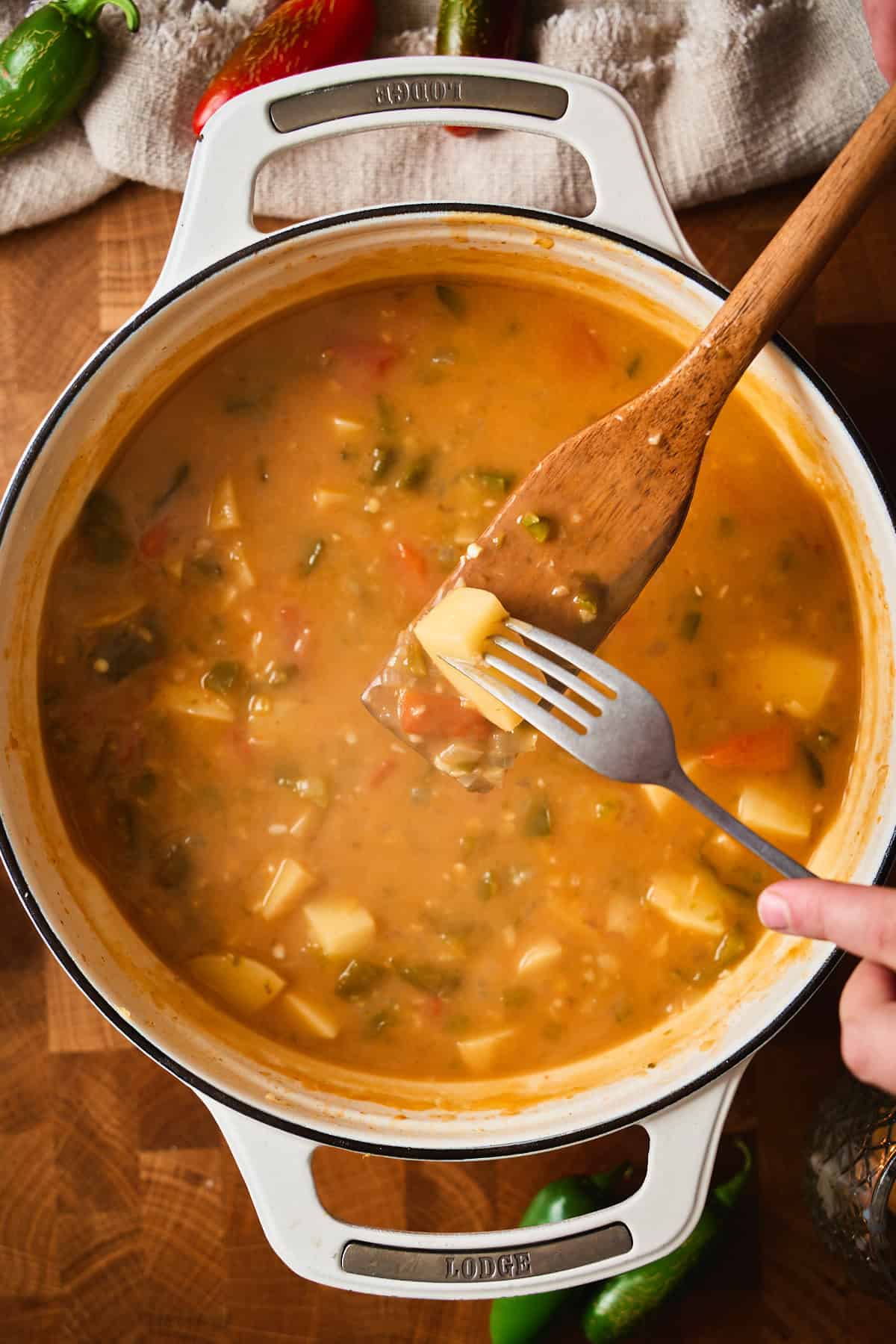 A fork testing a tender potato cube on a wooden spoon over a pot of jalapeño popper soup.