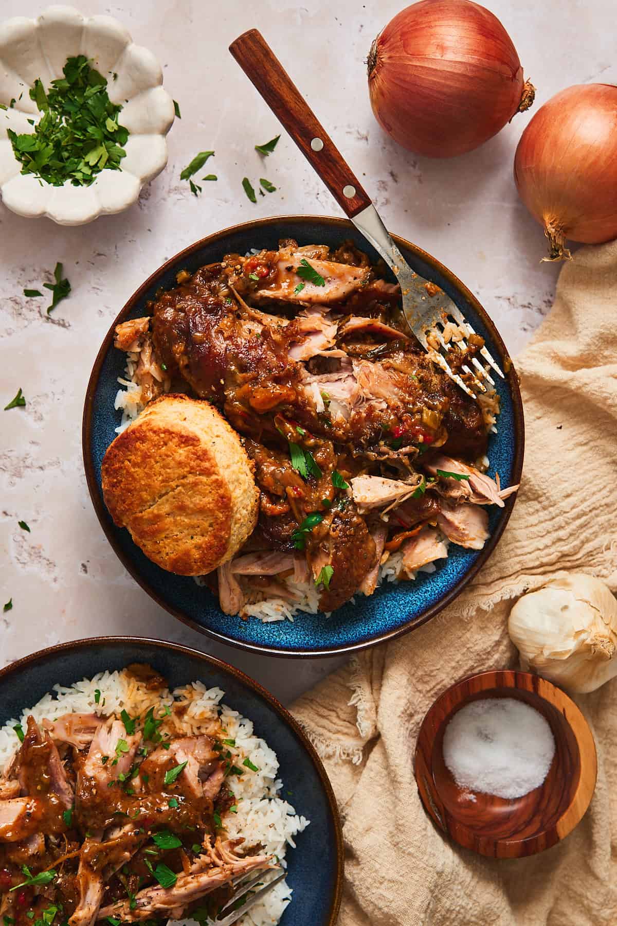 Smothered turkey wings in gravy over white rice in a blue bowl with a cornbread biscuit and parsley.